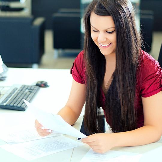 Junge Frau mit langen braunen Haaren liest lächelnd Unterlagen an einem modernen Büroschreibtisch.